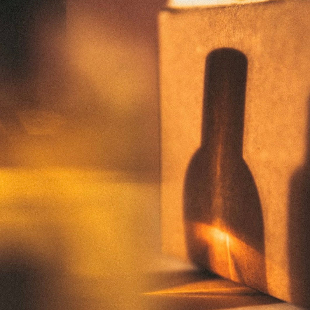 A warm, atmospheric close-up of a dark bottle-shaped object casting a shadow against a textured beige wall, illuminated by a golden light in a shallow depth of field.