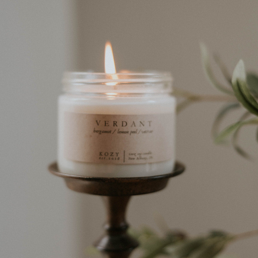 Candle in a jar labeled 'Verdant' on a wooden holder with a blurred plant in the background.