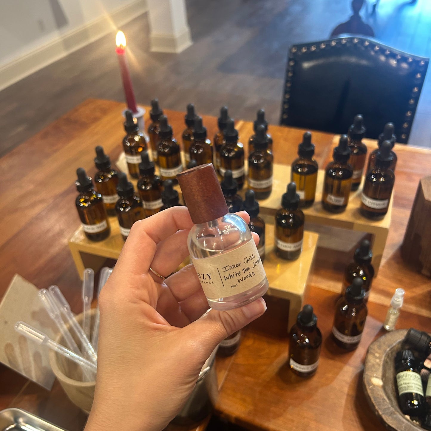 A hand holds a small clear glass bottle with a brown cap in a shop setting, with many amber dropper bottles displayed on a wooden counter in the background.