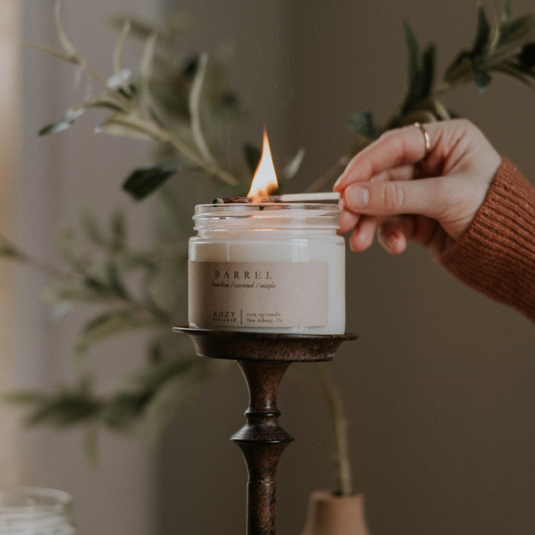 Hand lighting a candle with a plant and other candles in the background