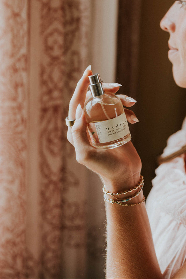 A woman in a white ruffled top holds a small glass perfume bottle with a silver spray nozzle, standing in profile near patterned pink curtains.