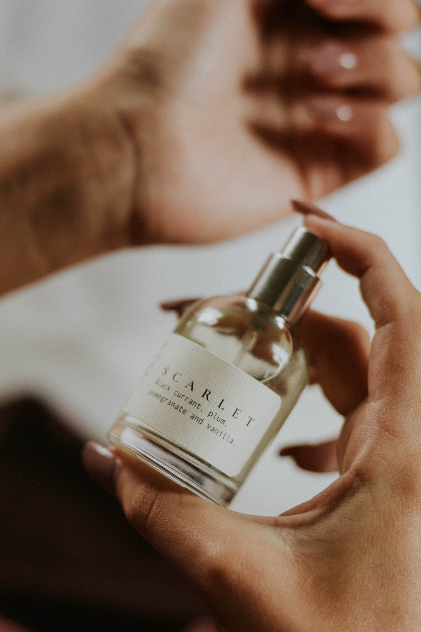 A person holding a small clear glass perfume bottle labeled SCARLET with a silver spray, while another hand holds a matching cap above, in a softly lit indoor setting.