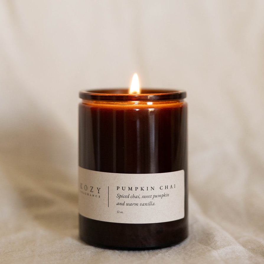 A lit amber glass jar candle with a white label reading 'KOZY CANDLE CO. PUMPKIN CHAI' on a soft beige fabric background.