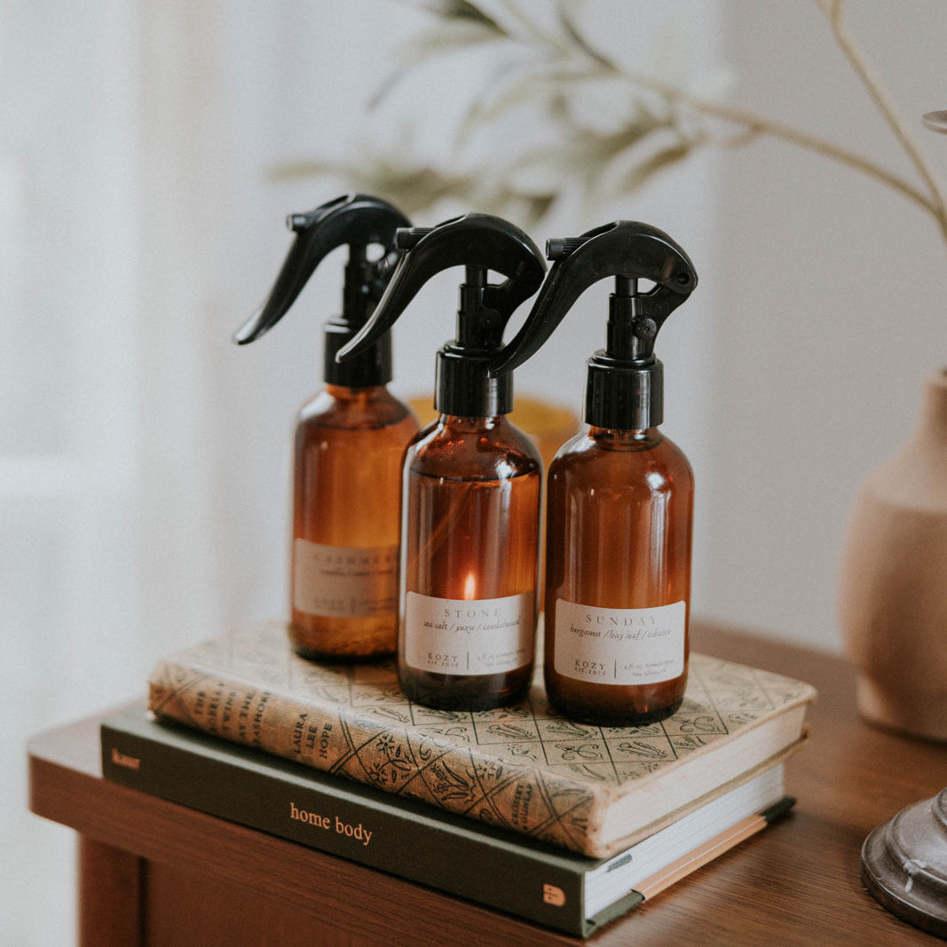 Three amber glass spray bottles with black nozzles on a stack of books, with a blurred plant in the background.