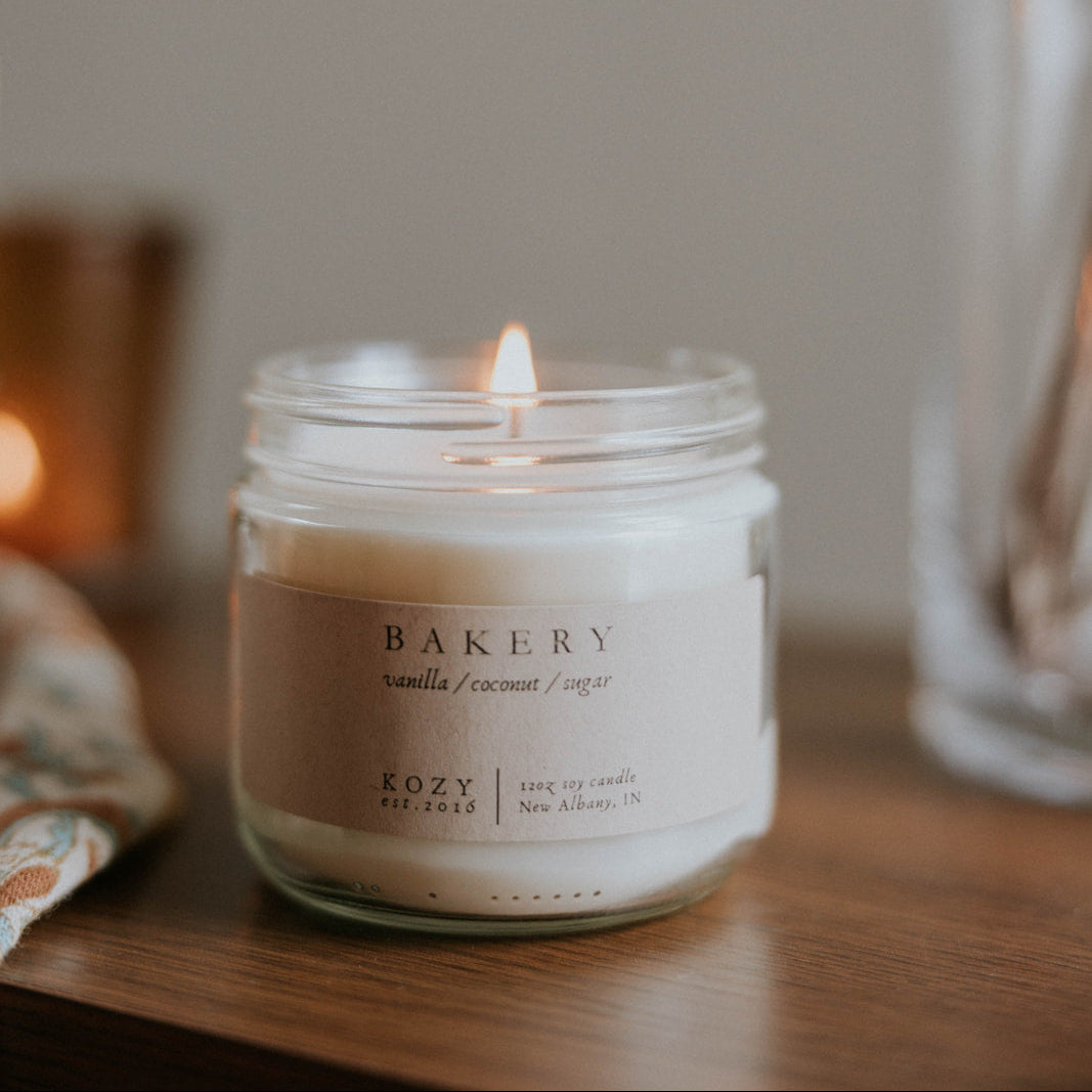 Candle in a jar labeled 'Bakery' on a wooden surface with a blurred background