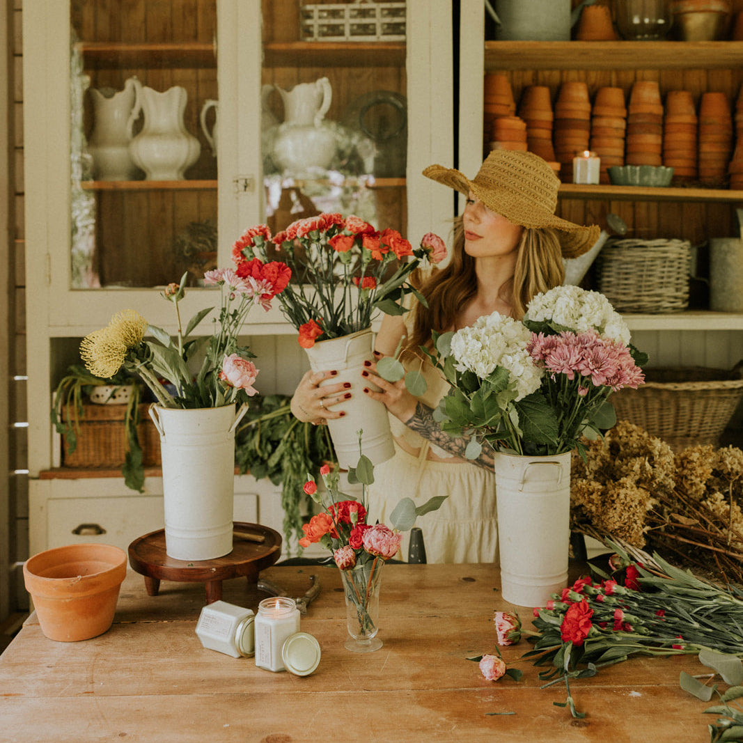 Woman arranging flowers in a rustic kitchen with wooden cabinets and shelves.