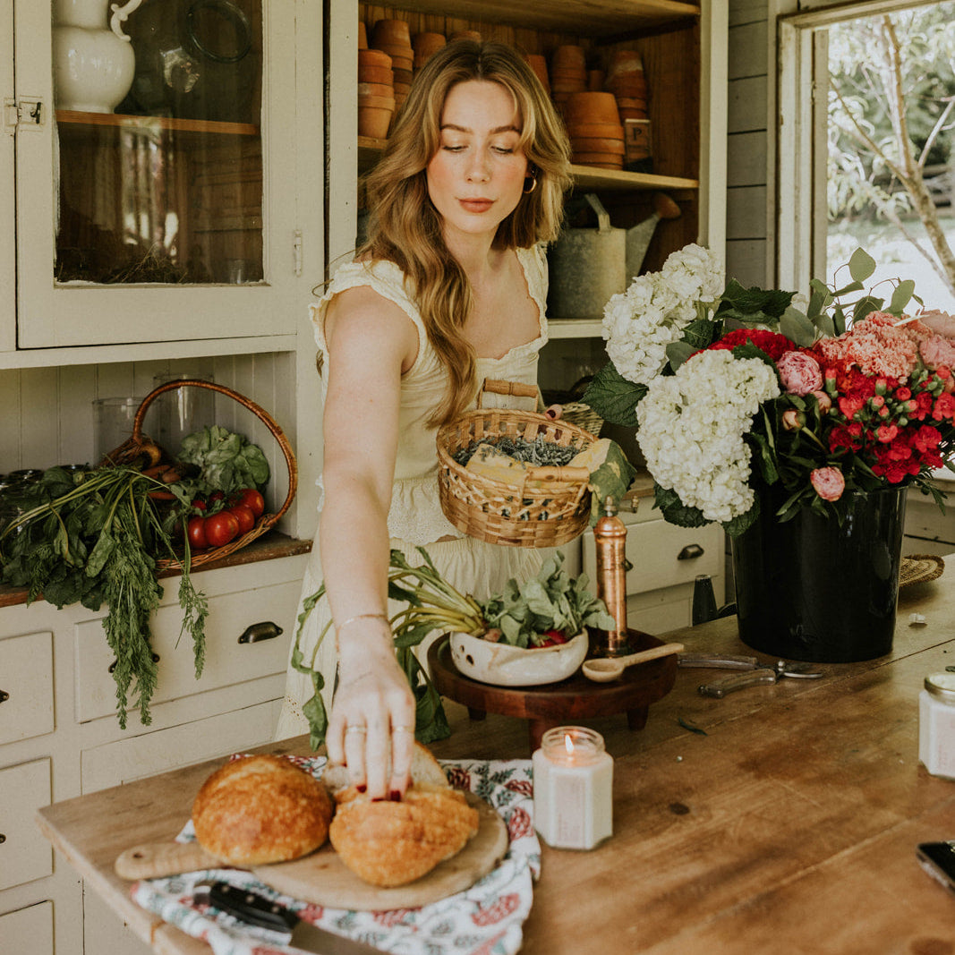 Woman preparing food in a rustic kitchen with flowers and bread on the counter.