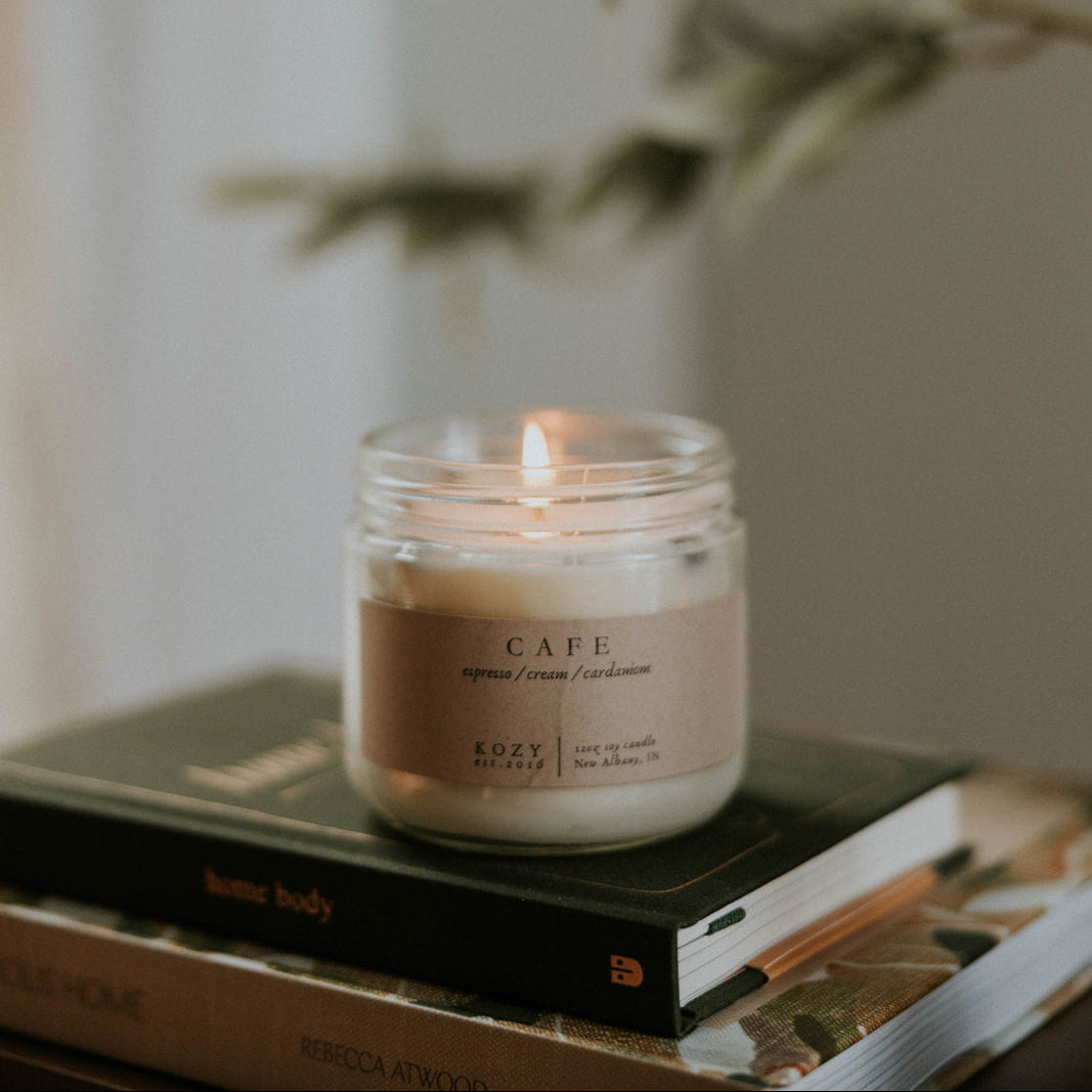 Candle in a glass jar scented 'Cafe' on top of books with a blurred plant in the background