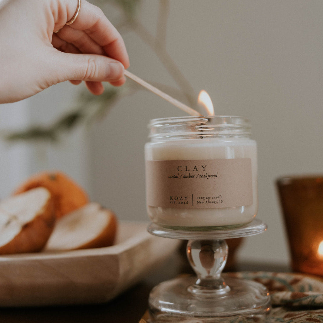 Person lighting a candle labeled 'CLAY' with a blurred background