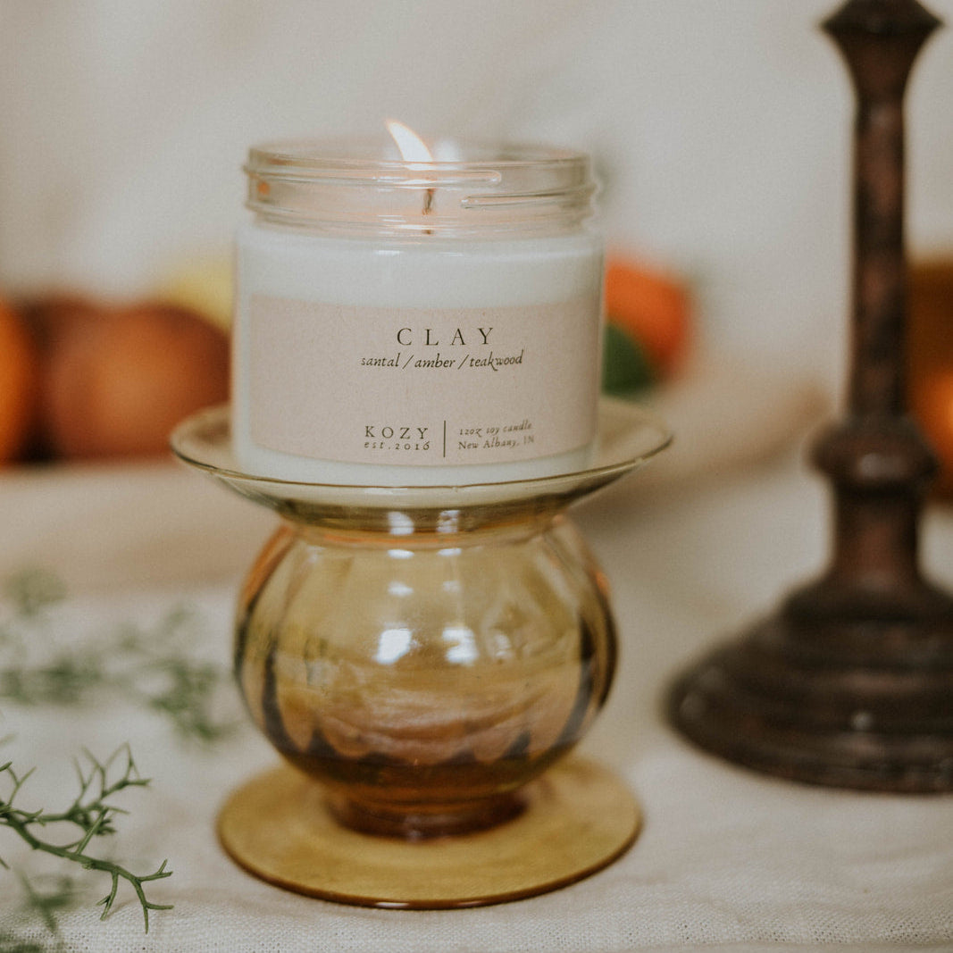 Candle in a glass jar labeled 'CLAY' on a decorative stand with a blurred background