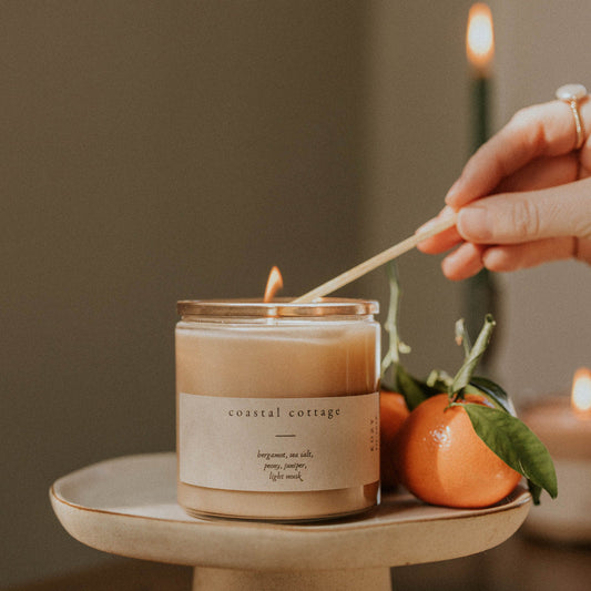Person lighting a candle labeled 'Coastal Cottage' on a small stone holder with fruits in the background.