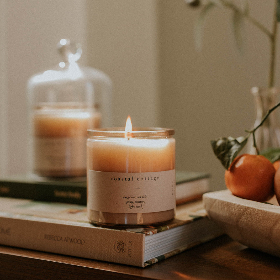 Candle with a label on a stack of books, surrounded by decorative items including a fruit and a plant.