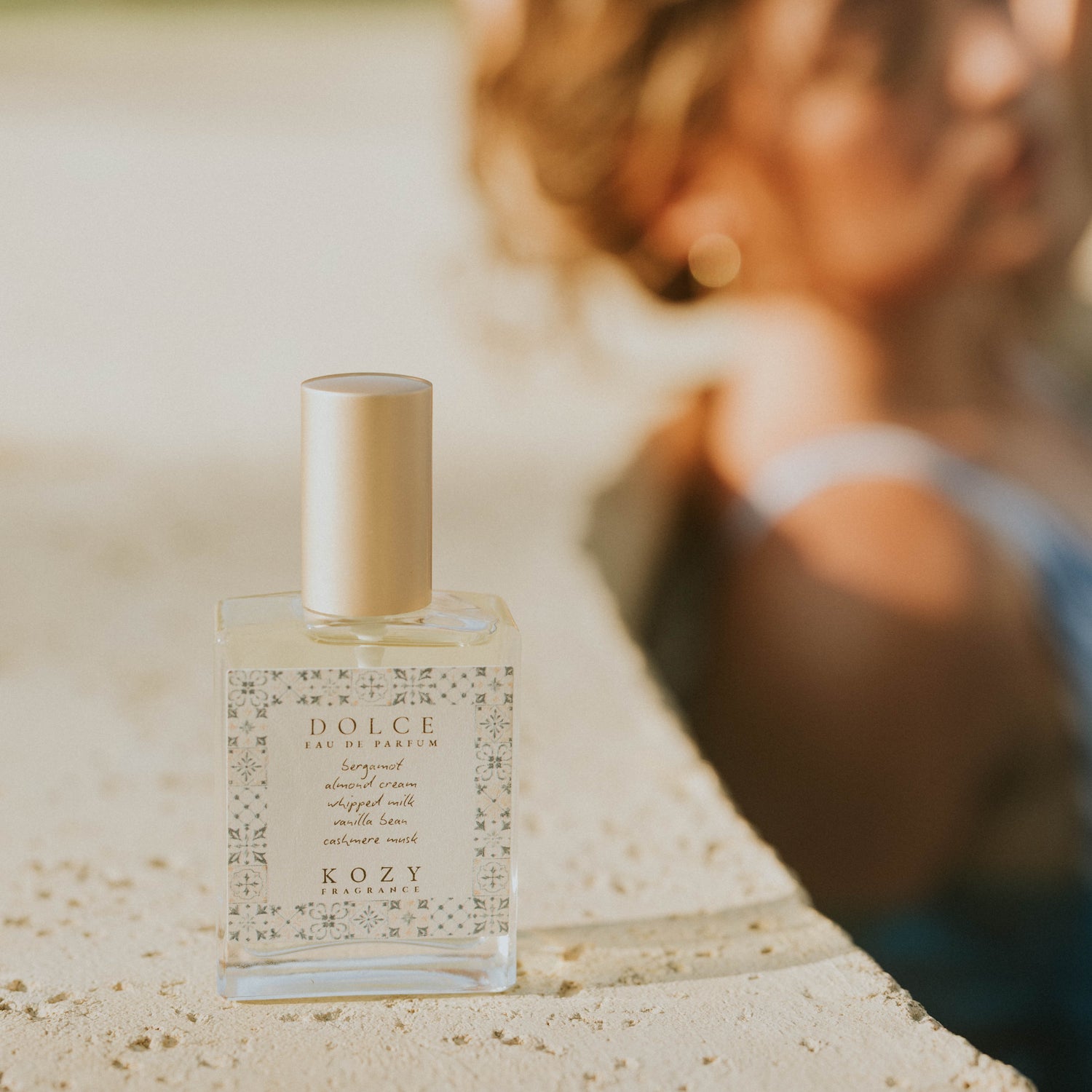 Dolce Kozy perfume bottle on a stone surface with a blurred person in the background.