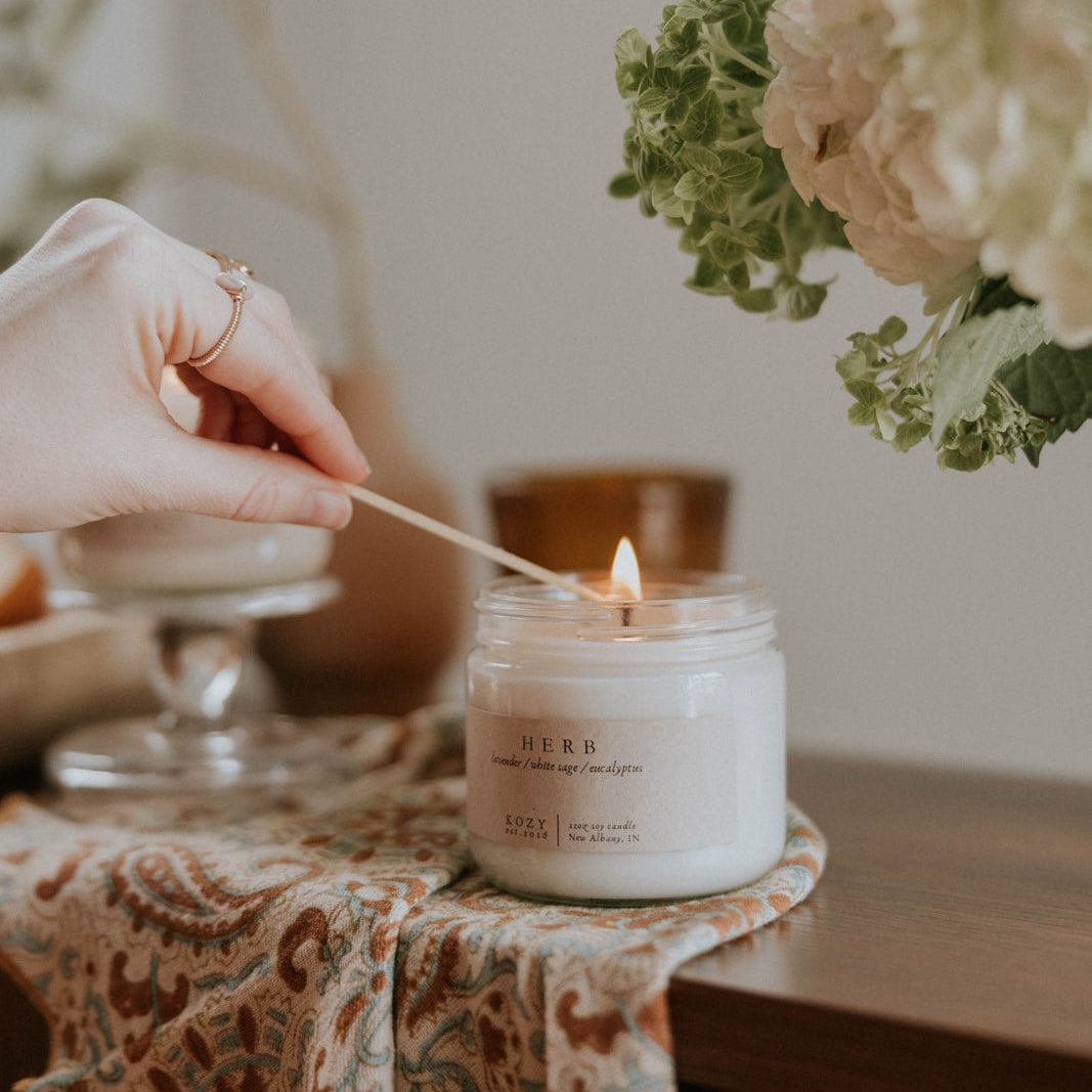 Person lighting a candle with a wooden wick, surrounded by flowers and a patterned cloth.