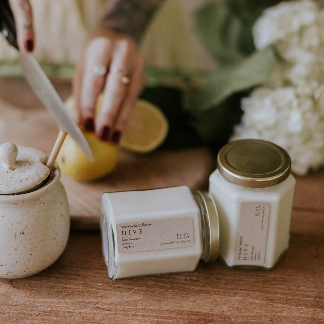 Person cutting a lemon with jars and a container on a wooden surface, surrounded by greenery and white flowers.