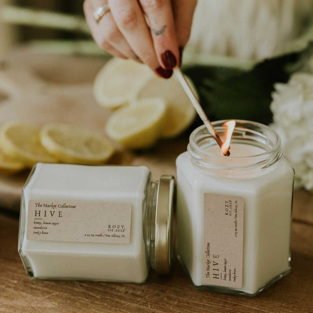 Person lighting a candle with a wooden wick, surrounded by lemons and flowers on a wooden surface.