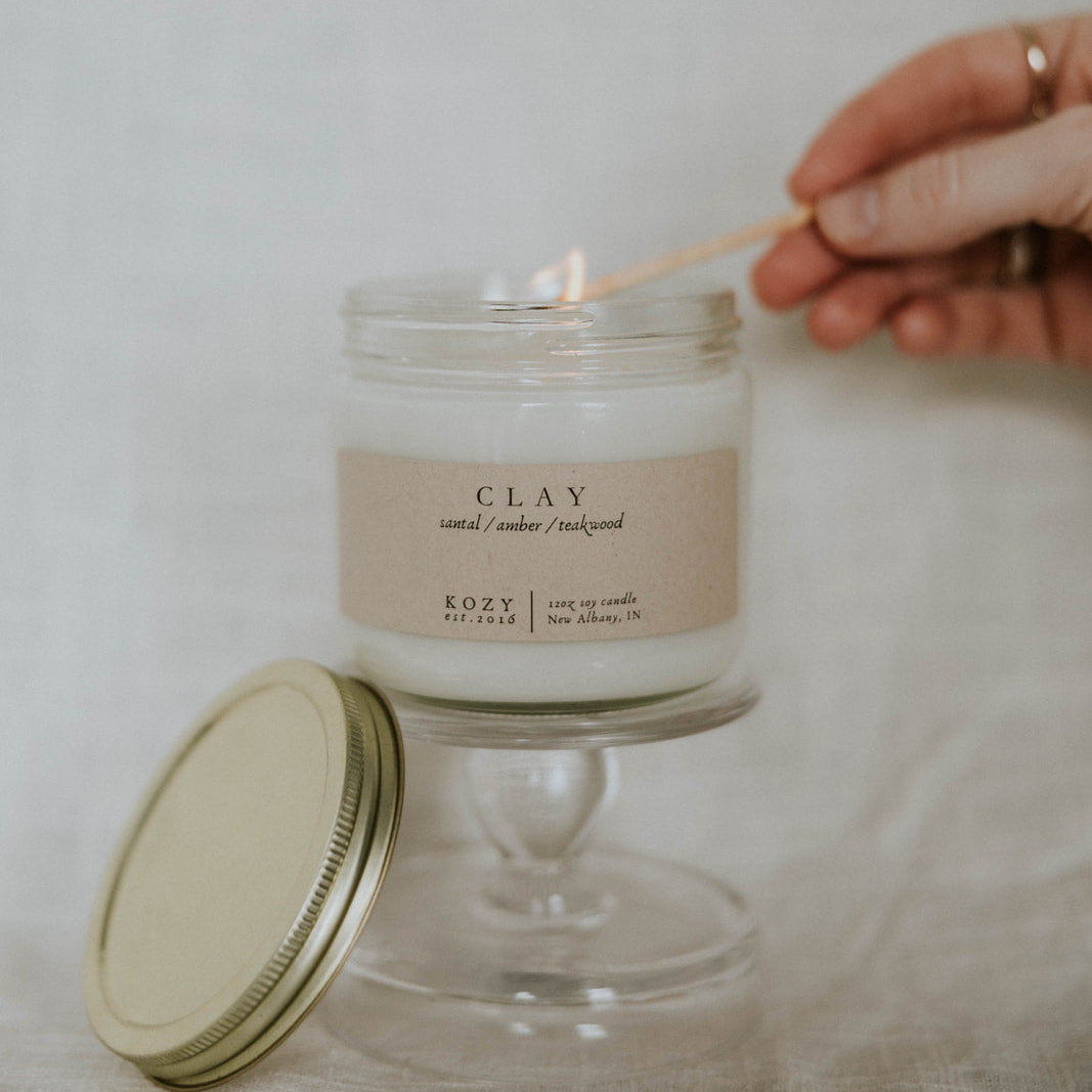 Candle being lit from a wooden wick holder with a 'Clay' candle jar on a neutral background