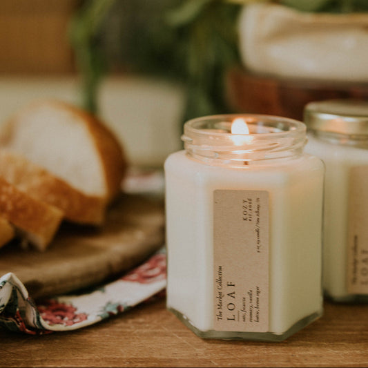 Candle with a label on a wooden surface with bread and greenery in the background, 'Loaf' scented