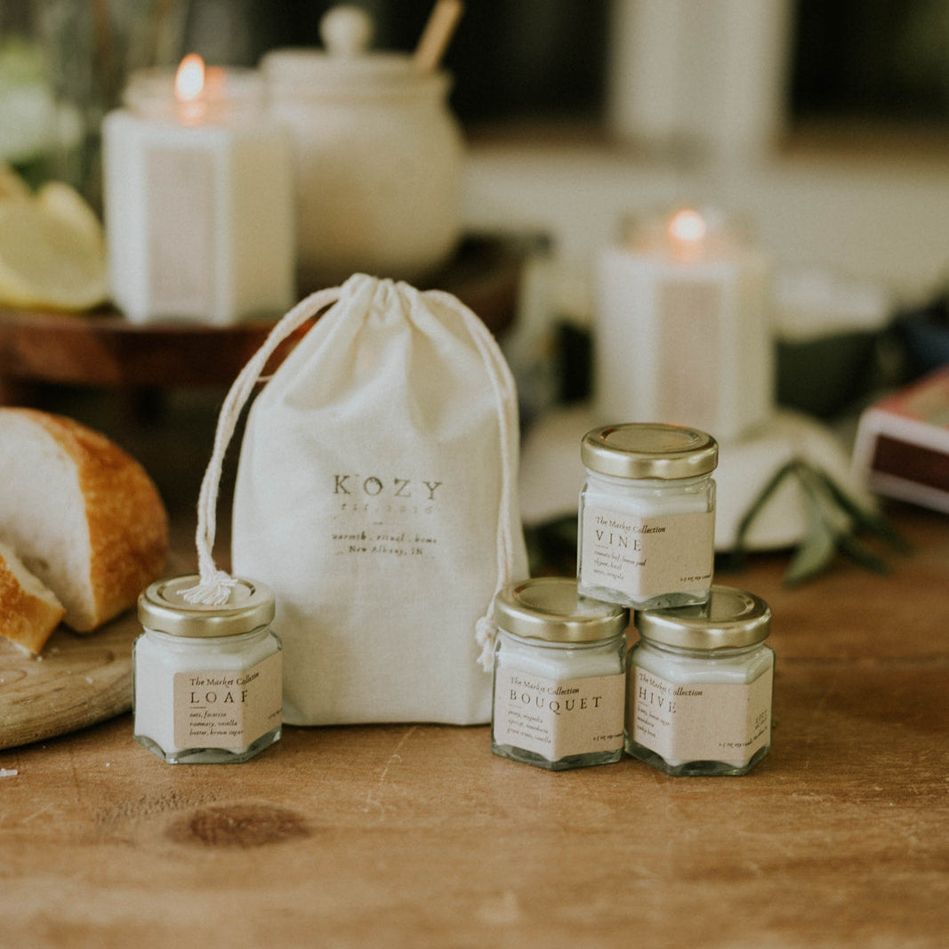 Kozy Kandles Market Collection on a wooden surface with a white drawstring bag and candles in the background.