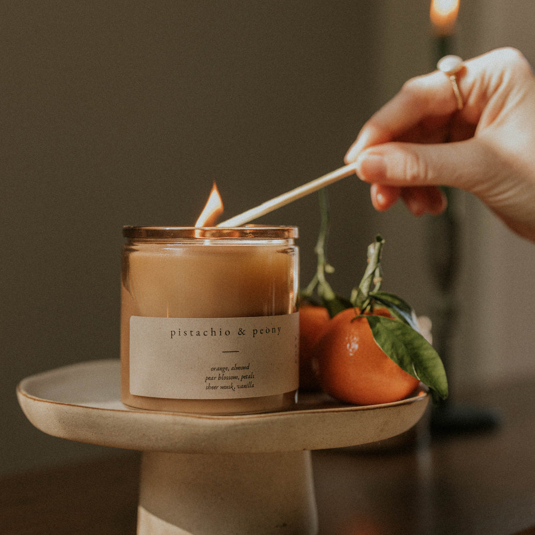 Person lighting a candle labeled 'Pistachio and Peony' on a wooden surface with a dark background.