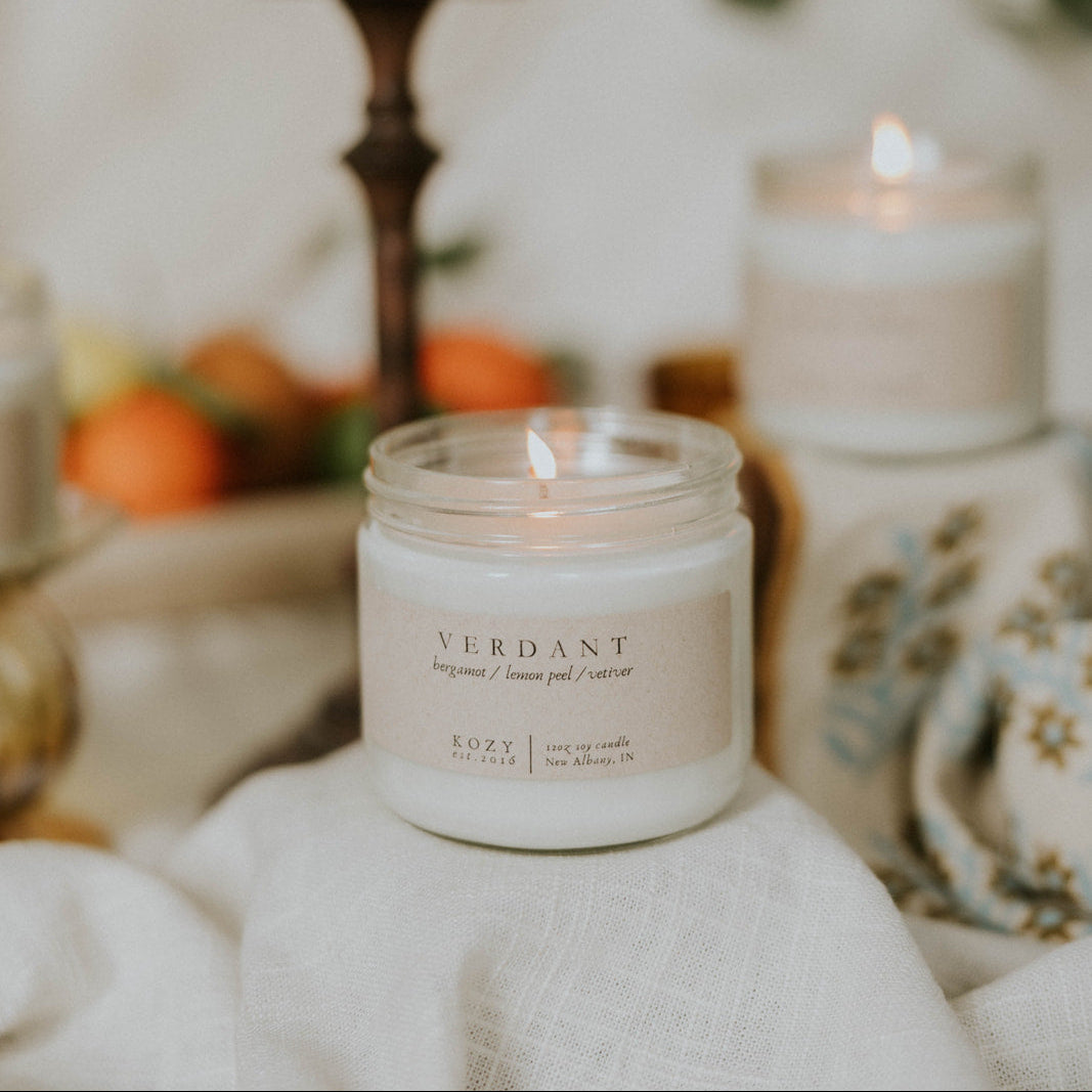 Candle scented 'Verdant' on a surface with other candles and decorative items in the background.