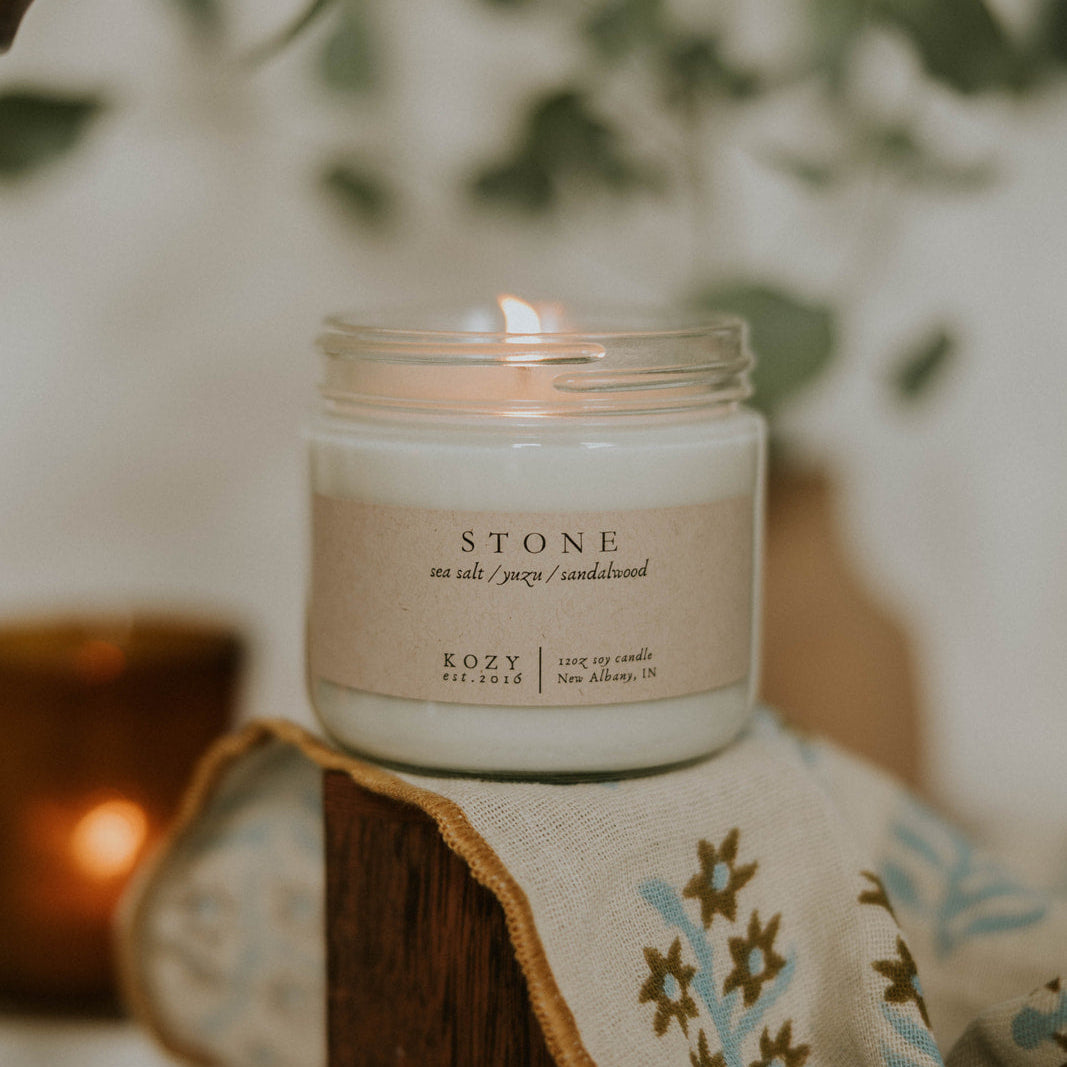 Candle in a glass jar labeled 'Stone' on a textured surface with a blurred background