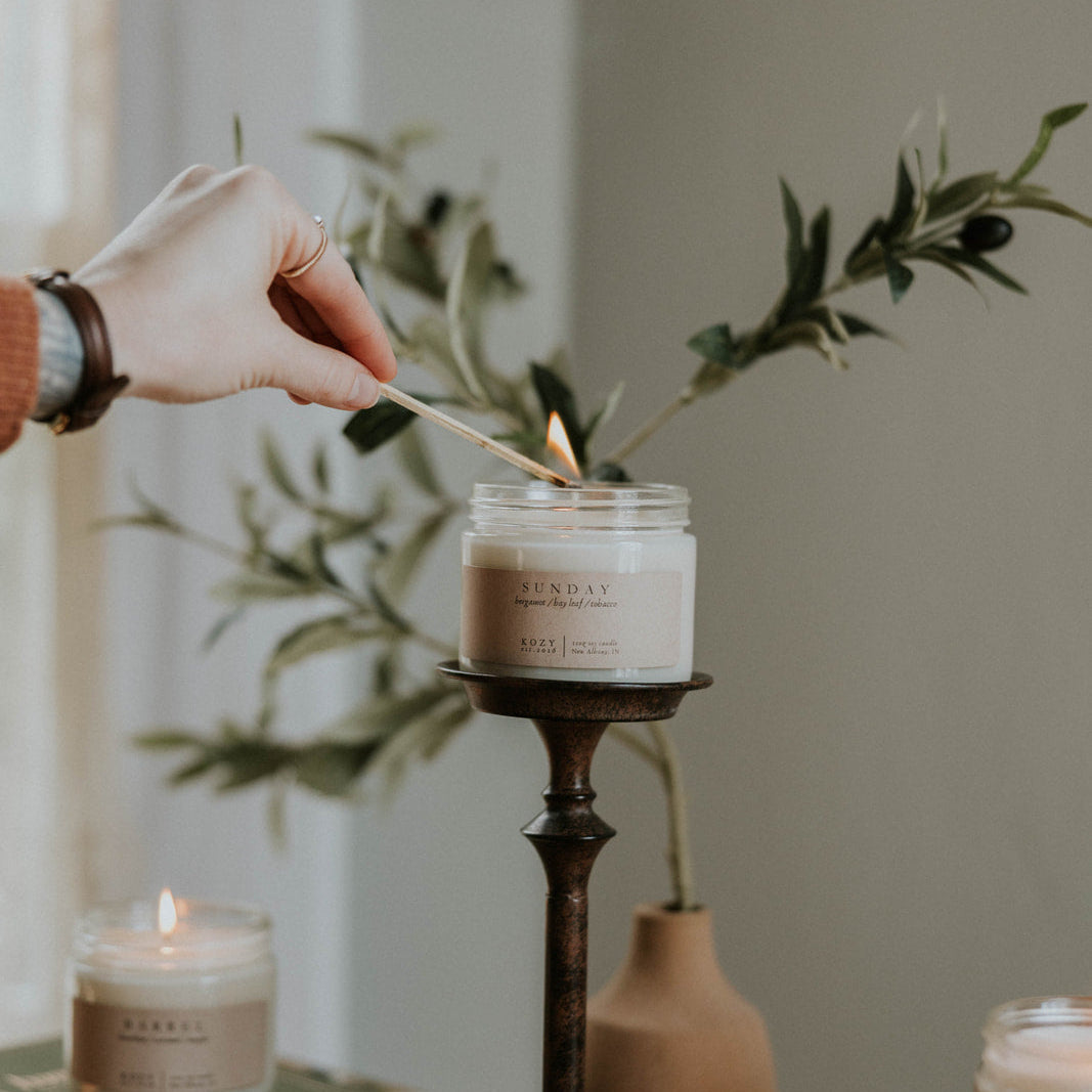 Person lighting a candle with a wooden spoon, surrounded by books and decorative items.