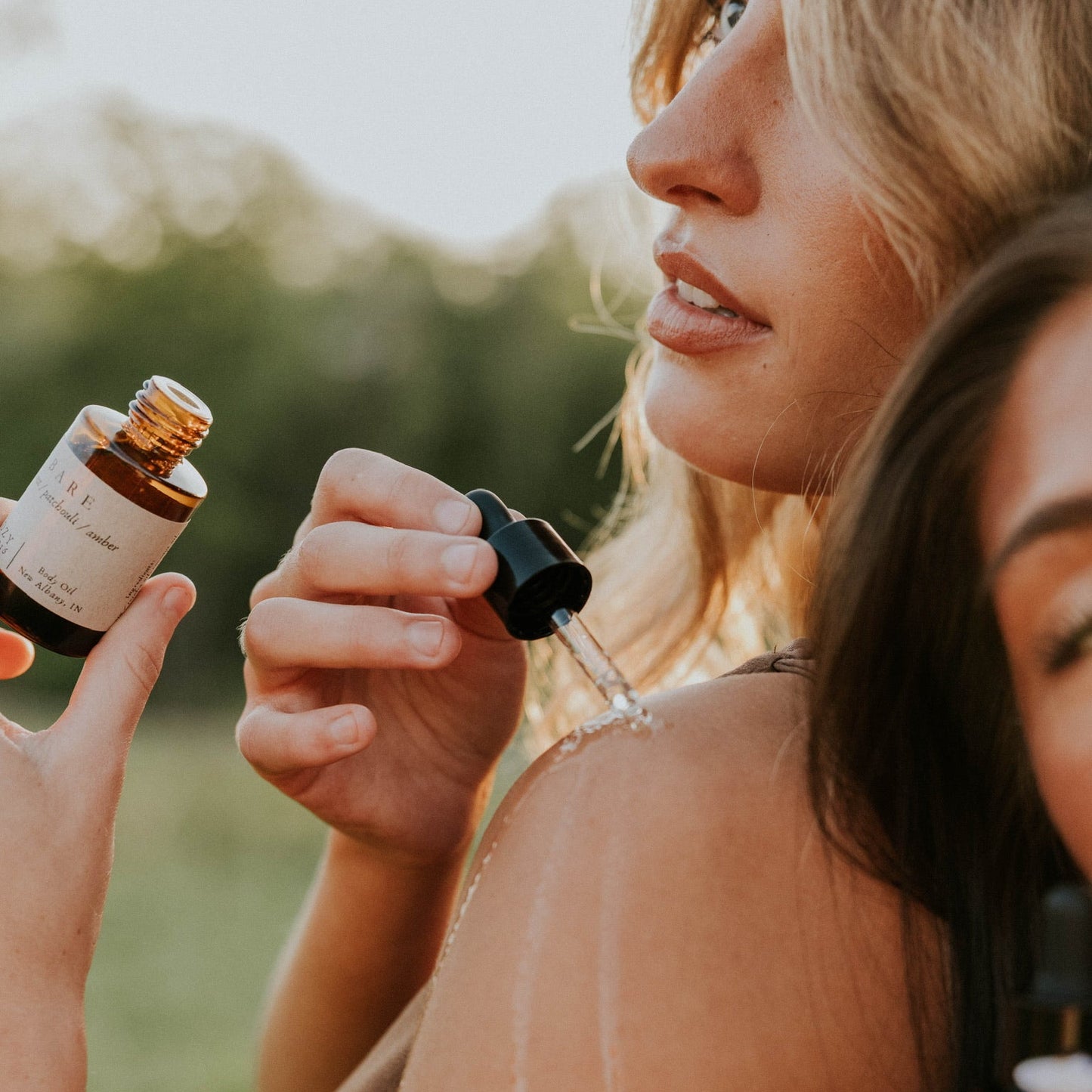 A woman outdoors, one pouring liquid from a dropper bottle on to her shoulder.