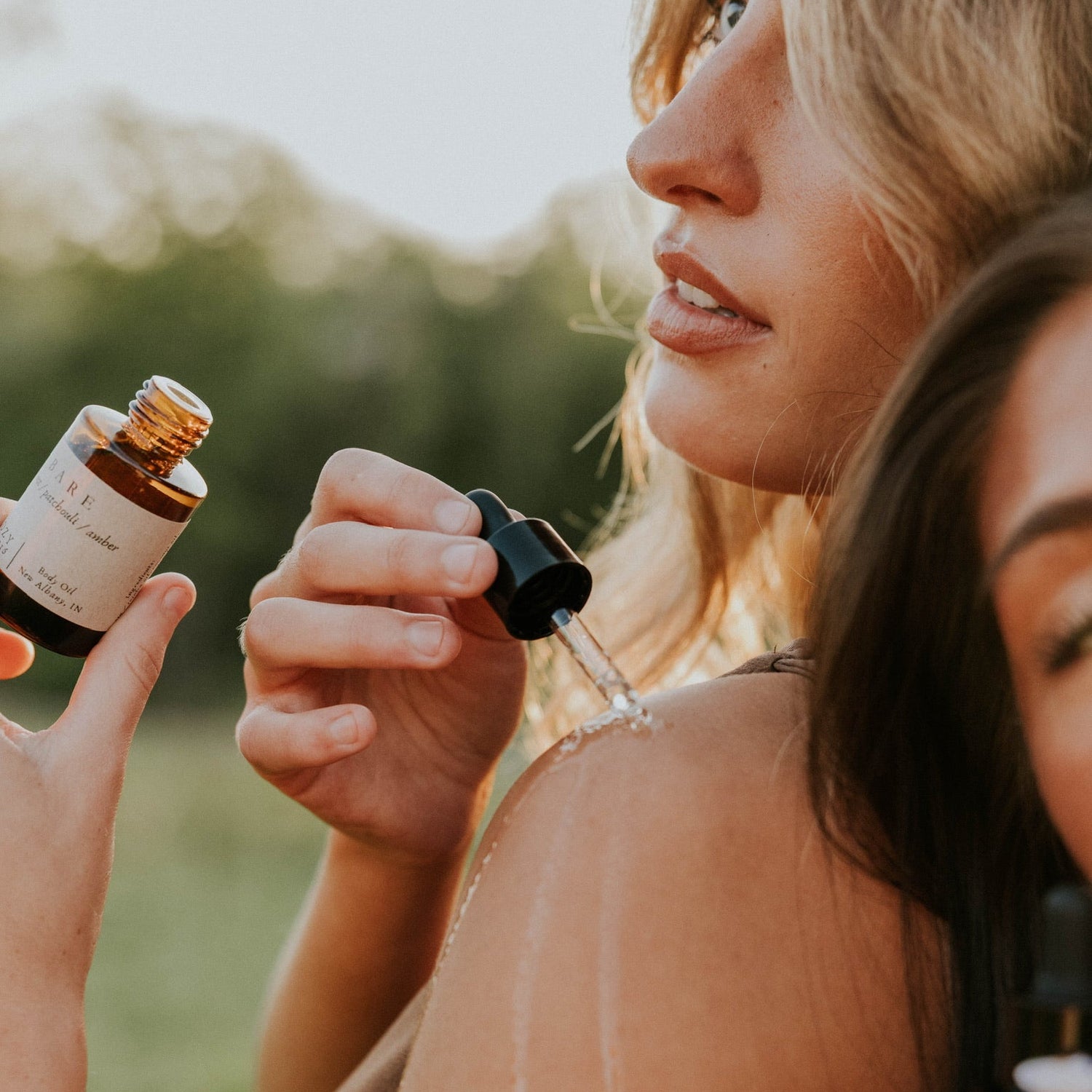 A woman outdoors, one pouring liquid from a dropper bottle on to her shoulder.