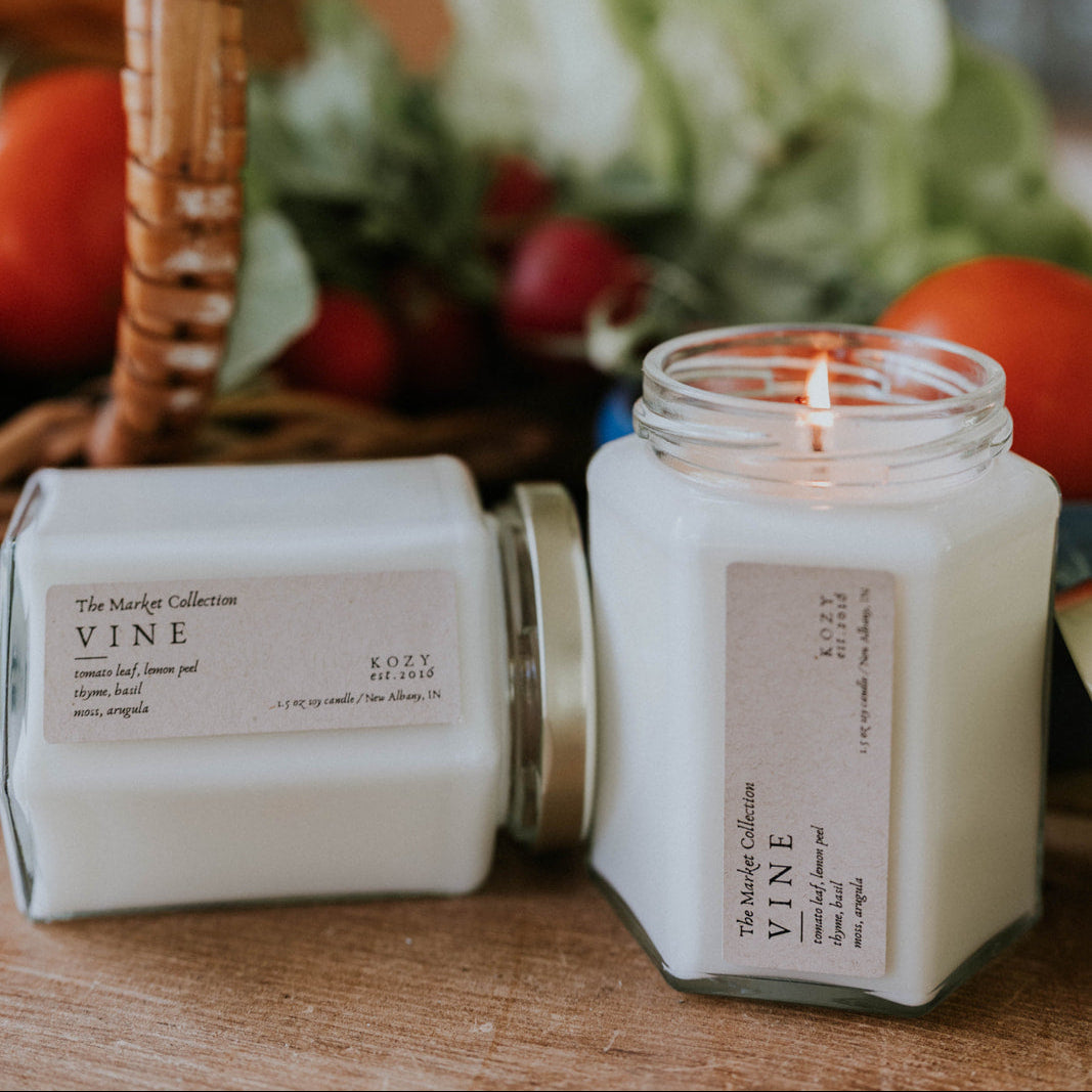 Candle and packaging scented 'Vine' on a wooden surface with a blurred background of fruits and flowers.