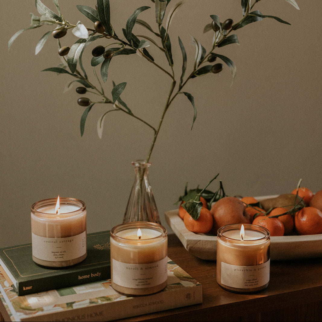 Three candles on a wooden surface with a plant and fruits in the background.