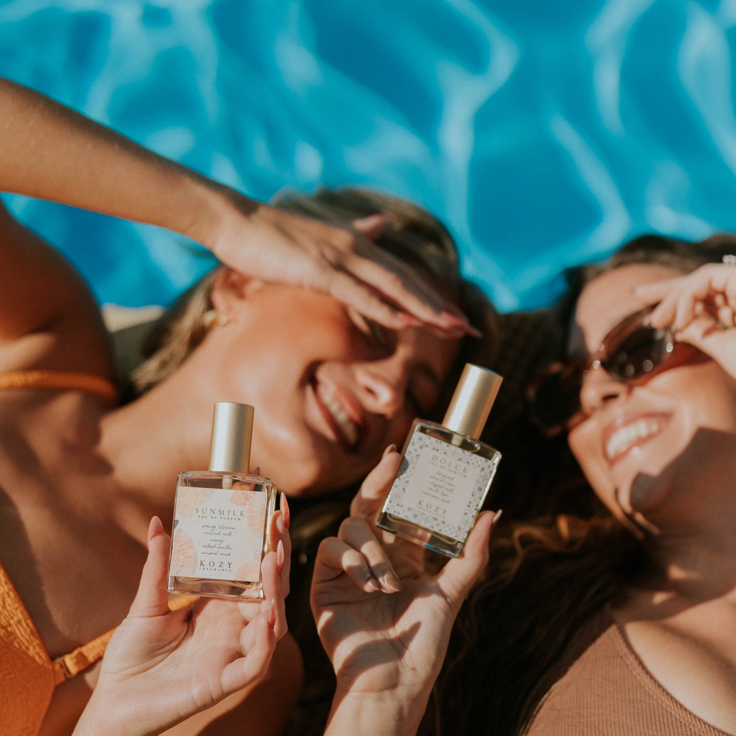 Two women by a pool holding perfume bottles.