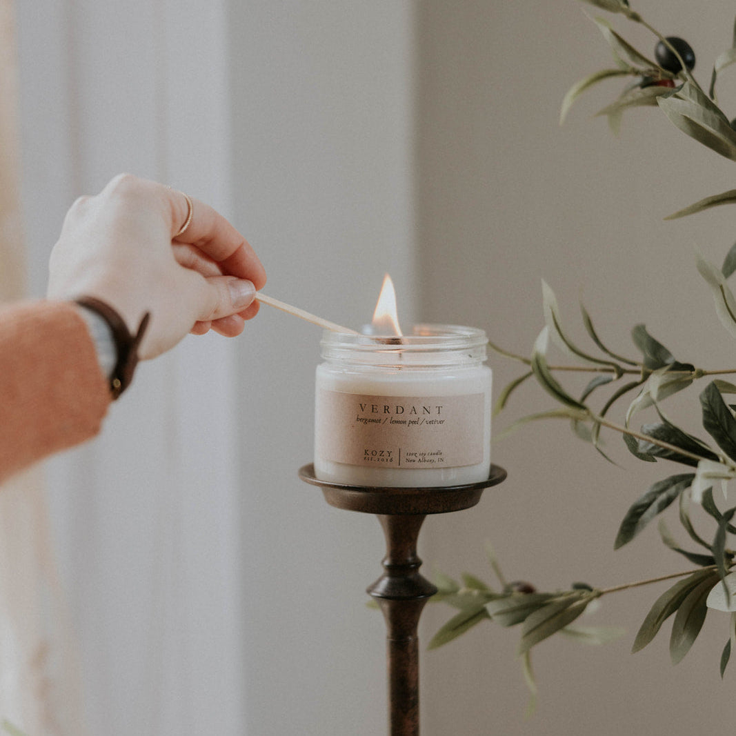 Person lighting a candle in a white container with 'Verdant' branding, surrounded by greenery.