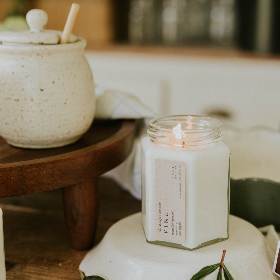 Candle in a glass jar on a white stand with green leaves, blurred background