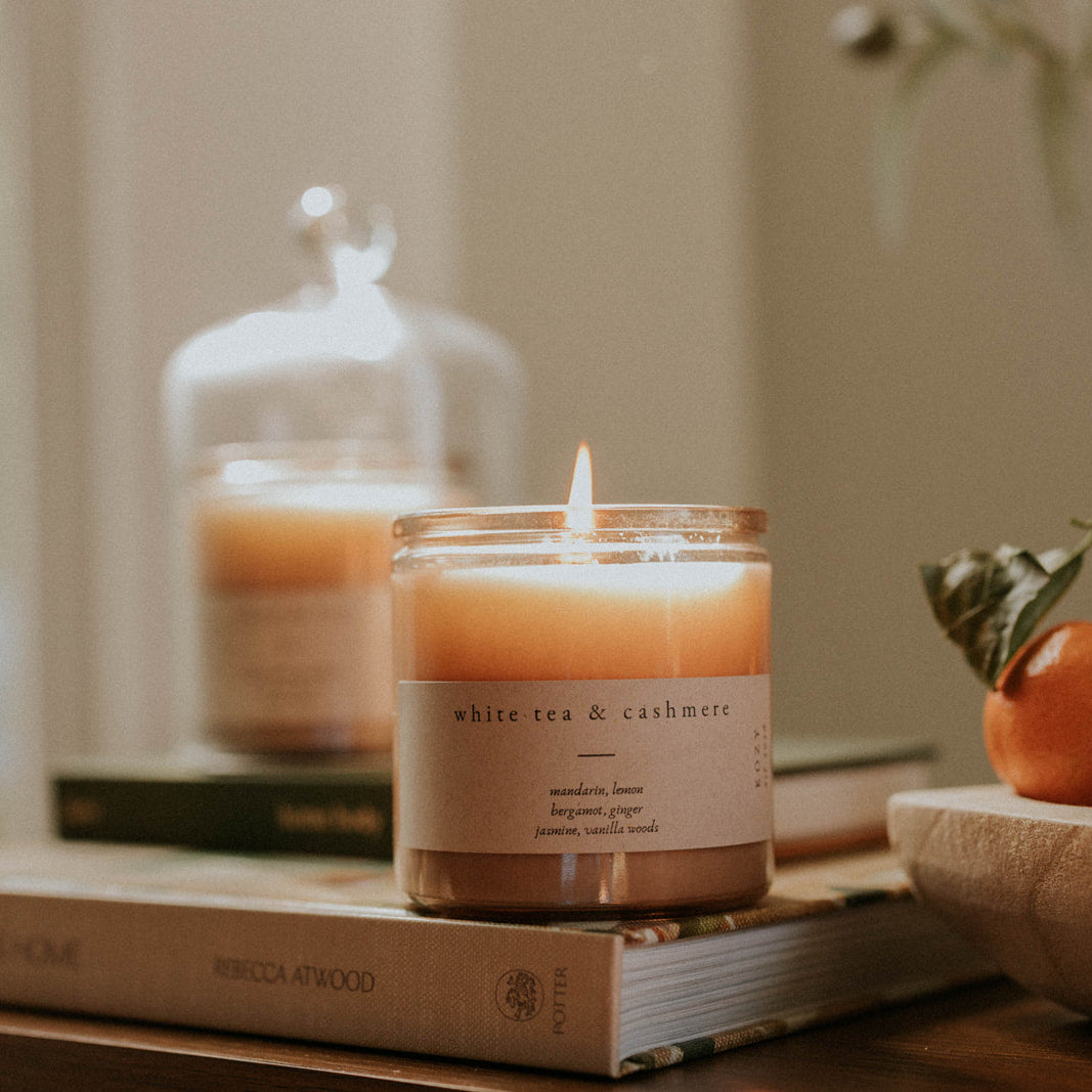 Candle with a label on a book, surrounded by fruits and a decorative jar.