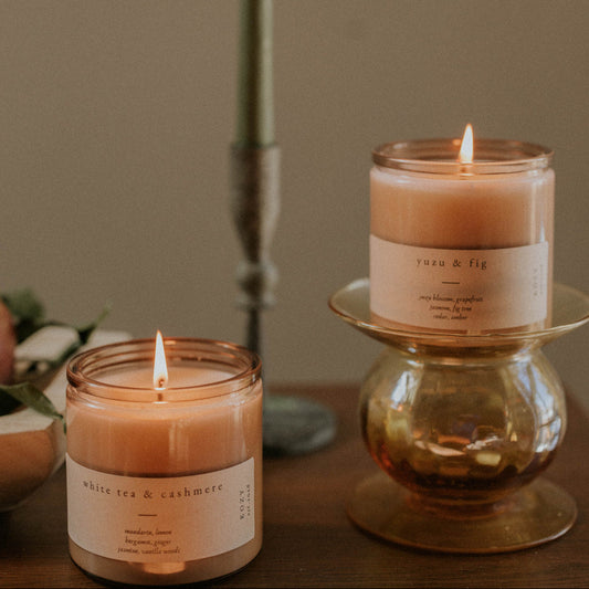 Two lit candles with visible labels on a wooden surface against a plain background