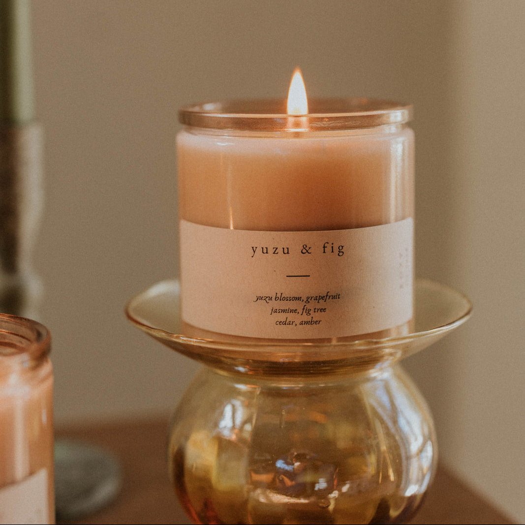 Candle labeled 'yuzu & fig' on a small glass stand with a blurred background