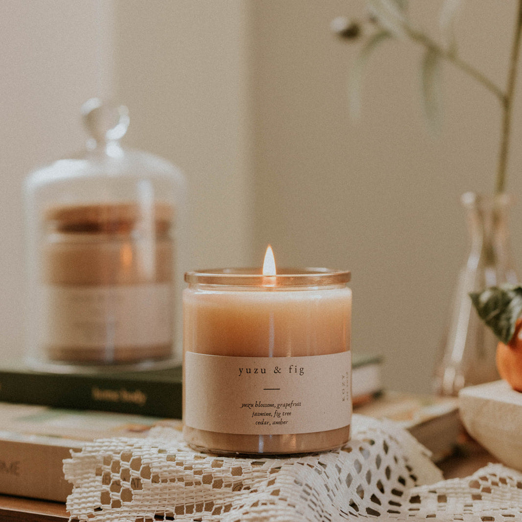 Lit Yuzu & Fig candle in a glass jar displayed on a lace cloth in a cozy home setting, creating a warm and relaxing ambiance.